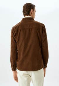 Camisa de pana marrón con un ajuste relajado, puños abotonados y cuello estándar. El material presenta rayas verticales y una textura suave.