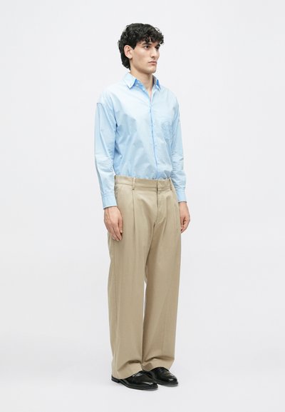 Šviesiai mėlyna marškinėlių uniforma su ilgomis rankovėmis, smėlio spalvos raukšlėti kelnes ir juodi batai. Modelis stovi aukštai prieš paprastą baltą foną.