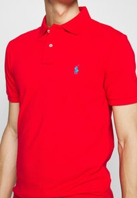 Röd bomullspolo med klassisk krage, treknapplåset och en blå broderad logga på bröstet. Korta ärmar, strukturerad tyg.