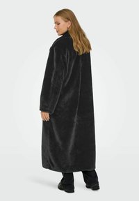 Donna con lunghi capelli biondi che indossa un lungo cappotto in pelliccia sintetica nera, pantaloni neri e scarpe nere, in piedi e con uno sguardo di lato.