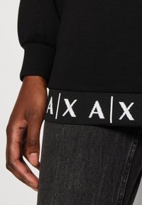 Černá mikina s balónovými rukávy, s kontrastním žebrovaným pasem, na kterém je bílé text "A|X A|X", a hladkým materiálem.