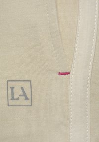 Beige stof met een gladde textuur heeft een grijs "LA" logo, en een roze accentsteek zichtbaar nabij de rand, die het ontwerp benadrukt.