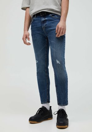 Man draagt versleten slim-fit blauwe spijkerbroek, witte sokken, zwarte sneakers met rubberen zolen en een grijs T-shirt tegen een effen achtergrond.