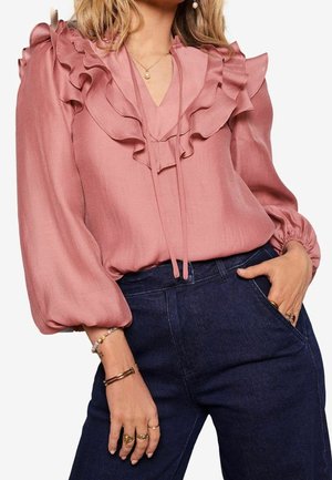Vrouw draagt een stofroze blouse met ruches en lange pofmouwen, gecombineerd met donkere, hooggesneden jeans en meerdere gouden ringen.