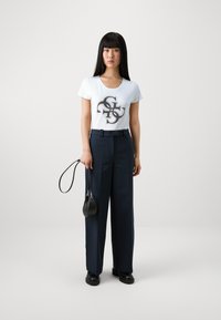 Camiseta blanca con un diseño gráfico negro, combinada con unos pantalones de pierna ancha de talle alto en color azul marino. La modelo sostiene un pequeño bolso negro.