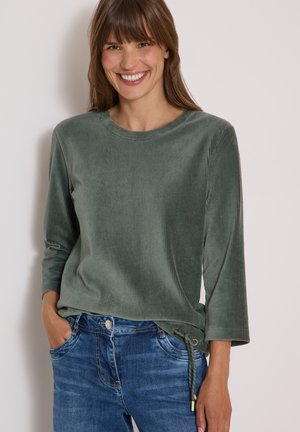Ung kvinde, der smiler, iført en grøn langærmet fløjlsbluse og blå jeans, stående med den ene hånd i lommen mod en ensfarvet baggrund.