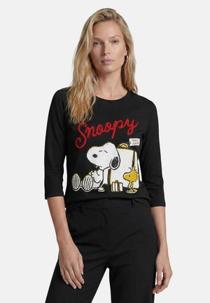 Blonde Frau trägt ein schwarzes Top mit Snoopy und Woodstock, sitzt neben einem Briefkasten mit dem roten Wort "Snoopy" darüber.