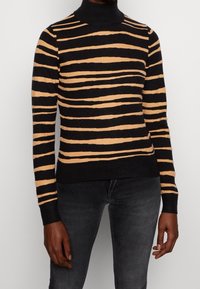 Pull à col roulé rayé noir et beige en maille côtelée, avec des lignes horizontales et des manches ajustées, associé à un jean en denim foncé.