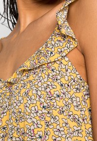 Blouse jaune à fleurs avec bretelles volantées, ornée de fleurs blanches accentuées de rose et de bleu, confectionnée dans un tissu léger et texturé.