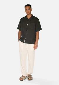 Schwarzes kurzärmliges Hemd mit Knopfleiste und Brusttasche, kombiniert mit hellbeigen Leinenhosen und grauen Sandalen. Minimalistisches Design und glatte Textur.