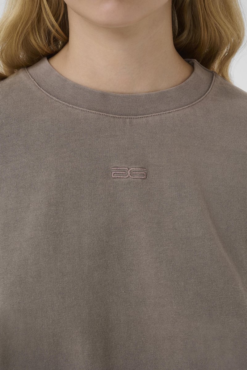Gros plan d'une personne portant un t-shirt col rond taupe avec un petit "26" brodé en fil assorti au centre de la poitrine.