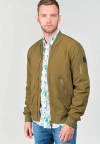 Olijfgroene bomberjack met een frontzipper, ribgebreide manchetten en een patch op de mouw. Draagt over een bloemenoverhemd met knopen.