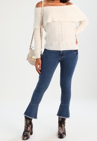 Cremfarbener Strickpullover mit schulterfreiem Design und geripptem Muster, kombiniert mit ausgestellten Jeans. Die Stiefel haben ein florales Muster und einen hohen Absatz.