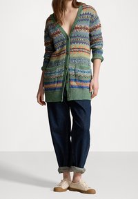 Grön mönstrad kofta med djup V-ringning, två fickor fram och träknappar. Bärs över mörkblå uppvikta jeans och beigea sneakers.