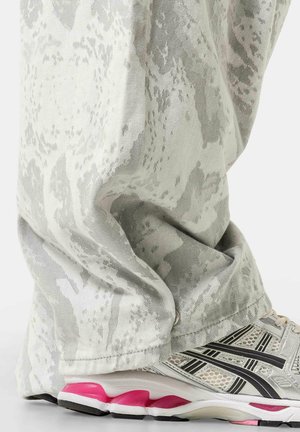 Pantalones con estampado blanco y gris cubriendo los tobillos sobre zapatillas deportivas blancas y rosas sobre un fondo liso.