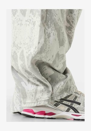 Pantalon blanc et gris à motif couvrant les chevilles au-dessus de baskets de sport blanches et roses sur un fond uni.