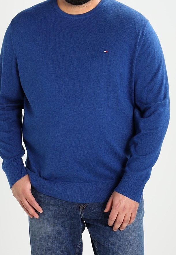 Homme portant un pull bleu à manches longues et un jean bleu, debout les mains près de la taille contre un fond uni.