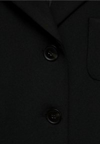 Blazer noir présentant une texture lisse, des revers crantés classiques, deux boutons noirs et un détail de poche poitrine.