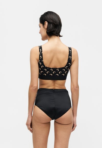 Γυναίκα με κοντά σκούρα μαλλιά που φοράει μαύρα ψηλόμεσα κάτω ρούχα και μαύρο crop top με μπεζ σχέδια ημισελήνου, γυρισμένη από πίσω.