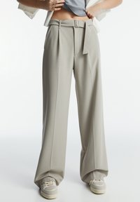 Hoge taille, wijde beige broek met riem, gecombineerd met beige sneakers, gedragen door een persoon met handen op de heupen tegen een effen achtergrond.