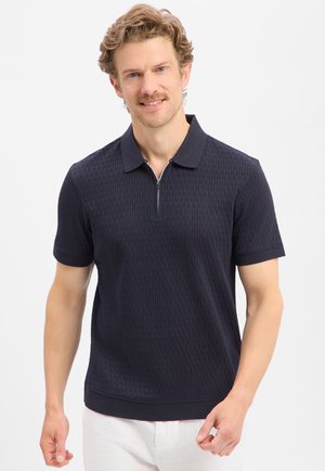 Homme aux cheveux bouclés châtain clair, portant un polo texturé bleu marine à manches courtes avec un col à fermeture éclair et un pantalon blanc sur fond blanc.