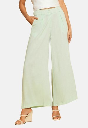Femme portant un pantalon vert clair à jambes larges avec des plis à l'avant et des sandales compensées beige avec des brides en corde, debout avec une main dans la poche.