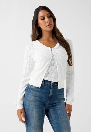 Femme portant un cardigan en maille blanc avec des boutons en perle par-dessus un top assorti et un jean bleu, posant devant un fond blanc.