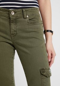 Pantalones cargo de color verde oliva fabricados con un tejido duradero, que cuentan con bolsillos frontales, un bolsillo lateral cargo y herrajes de botón plateados.