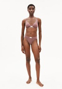 Vrouw die blootsvoets staat in een mauve tweedelige ondergoedset met merklabels op de bh en slip tegen een effen witte achtergrond.