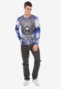 Langärmliges blaues Batik-Shirt mit Grafikdruck, das ein Totenschädel und Flügel zeigt. Kombiniert mit schwarzen Jeans und weißen Sneakers.