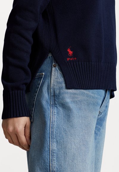 Marineblauwe gebreide trui met geribde mouwen en zoom, voorzien van een klein rood geborduurd logo. Gepaard met lichtblauwe spijkerbroek.