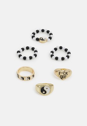 Cinco anéis com contas pretas e brancas, bandas de ouro e designs que incluem o símbolo do yin-yang e acentos de texto em cores contrastantes.