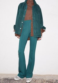 Cazadora de pana color verde azulado sobre un suéter de cuello alto marrón, combinada con pantalones acampanados verde azulado y zapatillas a rayas blanco y negro. Texturas suaves en toda la ropa.