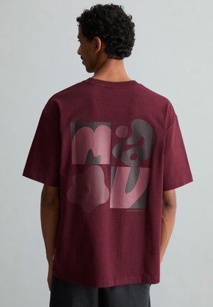 Man met krullend haar die een losse kastanjebruine t-shirt draagt met abstracte roze en grijze vormen op zijn rug, staand tegen een effen grijze achtergrond.