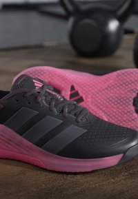 Μαύρα και ροζ αθλητικά παπούτσια Adidas σε ξύλινο πάτωμα με μια μαύρη αλτήρα (kettlebell) θολή στο φόντο.