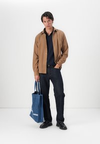 Une chemise marron à bouton sur un polo marine, un jean en denim foncé, des chaussures noires, et un sac fourre-tout bleu avec du texte blanc. Couleurs unies et design simple.