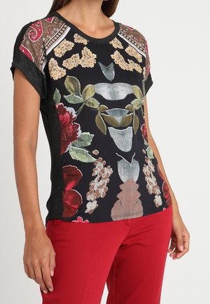 Kobieta ubrana w czarną bluzkę w kwiatowy wzór z krótkim rękawem, z akcentami czerwieni, beżu i zieleni, w połączeniu z jaskrawoczerwonymi spodniami.