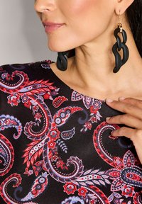 Schwarzes Kleid mit einem bunten Paisleymuster in Rot, Blau und Weiß. Große schwarze Kettenohrringe mit einem goldenen Akzent. Glatte Stofftextur.