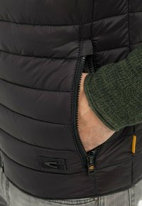 Mano en el bolsillo con cremallera de un chaleco acolchado negro, usado sobre un suéter de punto verde y pantalones grises.