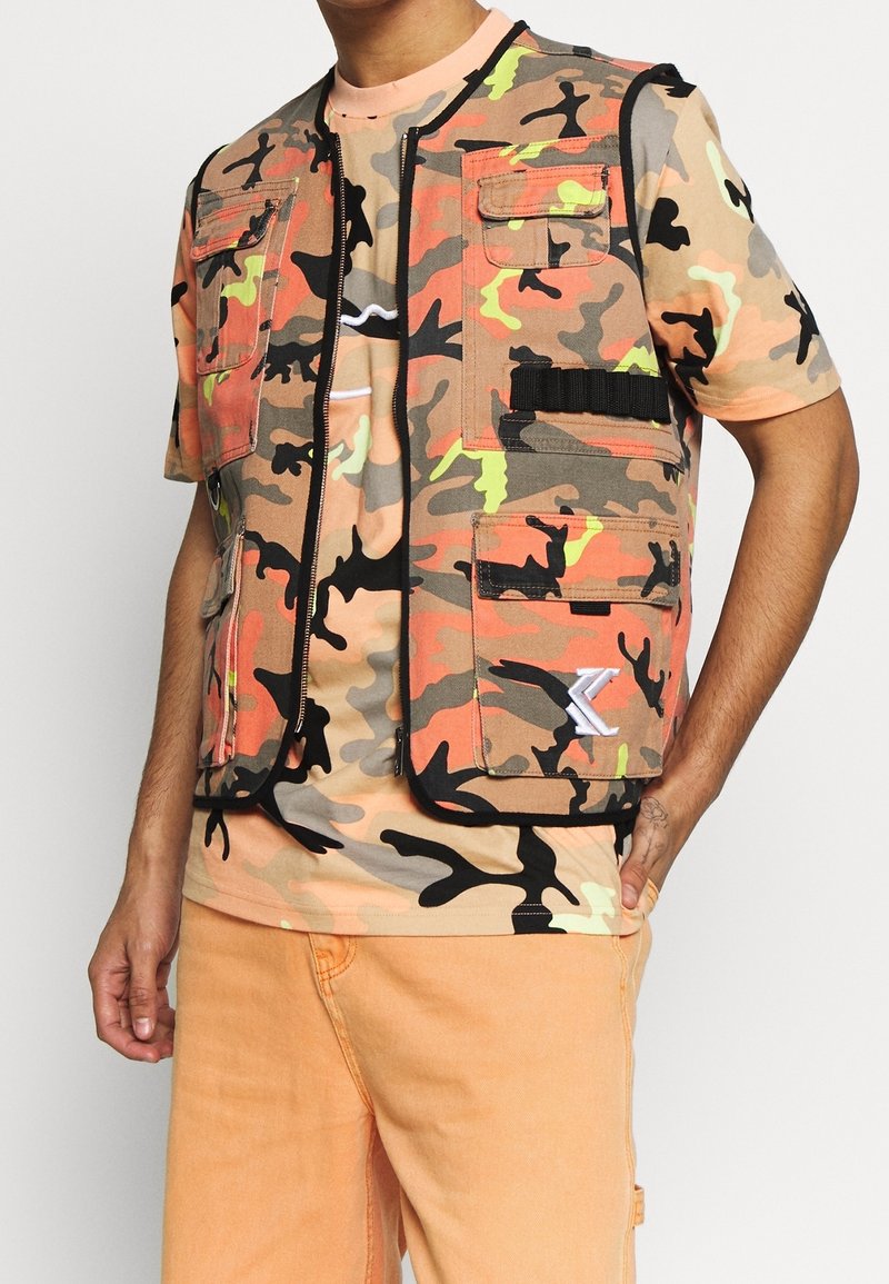 Homme portant un gilet camouflage orange à poches multiples sur un t-shirt camo assorti et un pantalon orange clair, main dans la poche.