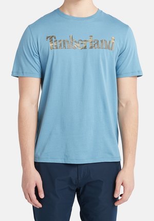 Homme portant un t-shirt Timberland bleu clair avec un logo camouflage sur la poitrine et un pantalon bleu foncé, debout devant un fond blanc.