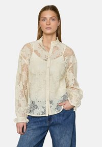 Kvinde iført en cremefarvet bluse med knapper og blonder, med flæsekrave og manchetter, kombineret med løse blå jeans, stående med den ene hånd i lommen.