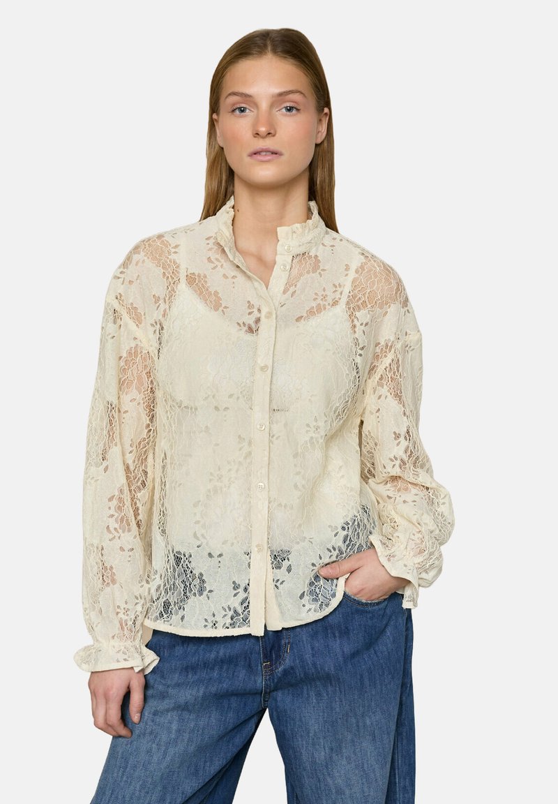 Kvinde iført en cremefarvet bluse med knapper og blonder, med flæsekrave og manchetter, kombineret med løse blå jeans, stående med den ene hånd i lommen.