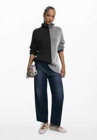 Pull en tricot à rayures verticales noires et blanches, col montant, associé à un pantalon en denim sombre ample et des chaussures de couleur claire. Tient un sac à motifs.