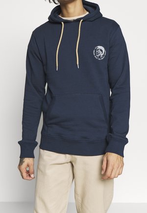 Homme portant un sweat à capuche bleu marine avec cordons beiges et petit logo sur la poitrine, associé à un pantalon beige clair, debout sur fond blanc.