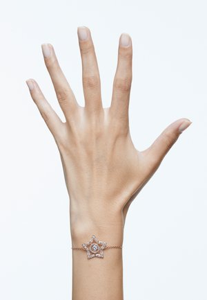 Bracelet en or rose avec un design d'étoile et un accent circulaire central, incrusté de petites pierres précieuses claires, porté sur une main.