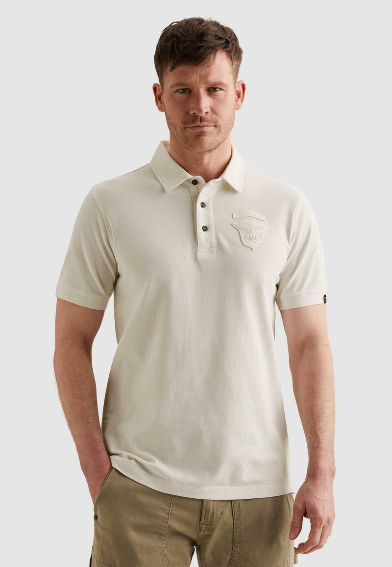 Homme aux cheveux courts portant un polo beige clair avec un écusson sur la poitrine et un pantalon kaki, debout avec une main dans la poche.