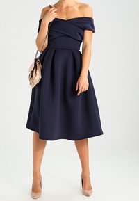 Robe bleu marine à épaules dénudées avec jupe plissée, corsage ajusté et tissu doux. Associée à un petit sac à main rose clair. Escarpins couleur neutre.