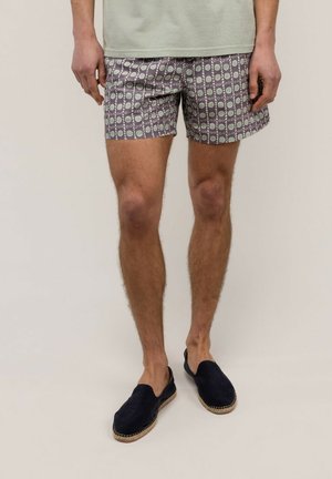 Hombre que lleva pantalones cortos estampados con círculos verdes y blancos, una camisa gris claro y zapatos negros sin cordones, de pie frente a un fondo liso.
