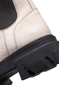 Bota sintética de color beige claro con una suela de goma negra texturizada. Presenta un empeine suave, panel elástico negro y logotipo en relieve.